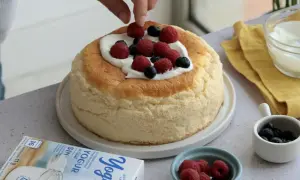 Tarta de queso esponjosa decorada con frutos rojos y nata. Se ve una caja de yogur griego al lado.