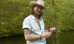 Brian Kelley, in a cowboy hat and light blue shirt, smiles while holding fishing line, perhaps inspired by his songs.