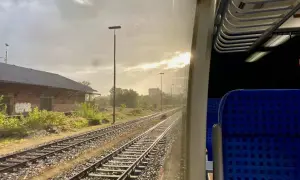 Blick aus dem Zugfenster auf Gleise und ein altes Backsteingebäude. Die Sonne bricht durch die Wolken. Man fragt sich, wie viel kostet eine Zugfahrt.