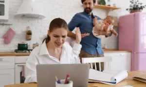 Zestresowana kobieta przy laptopie, mąż z dzieckiem w tle. Czy przerwa na karmienie piersią do jakiego wieku dziecka jest możliwa w takiej sytuacji?