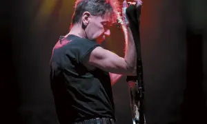 Muzyk w czerni, z czerwoną poświatą na włosach, przygotowuje się do wykonania swoich ksu utworów na gitarze.