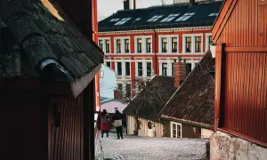 Två personer promenerar på en kullerstensgata i Oslo centrum, omgivna av charmiga trähus och en ståtlig röd byggnad.