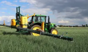 Zielony traktor z opryskiwaczem polowym, gotowy do pracy z adiuwantem, na tle pola i chmur.