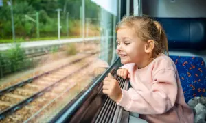 Uśmiechnięta dziewczynka wpatruje się przez okno pociągu. Zastanawia się, od ilu lat można jeździć samemu pociągiem, obserwując mijane krajobrazy.