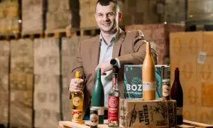 Mężczyzna w brązowej marynarce prezentuje butelki z alkoholem, w tym te z etykietą "Spice Bros". W tle magazyn pełen towarów, sugerujący dystrybucję california alkohol.