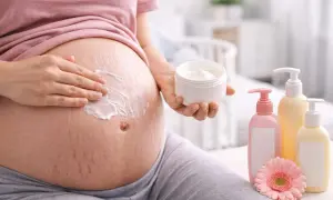 Ciężarna kobieta smaruje brzuch kremem. W pobliżu stoją butelki z kosmetykami i kwiat. Bezpieczne dla skóry w ciąży, np. bez kwasu glikolowego.