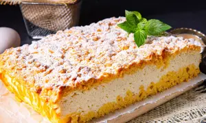 Puszysty sernik z kruszonką, posypany cukrem pudrem, ozdobiony listkiem mięty. Obok leżą jajka, sugerujące domowe wypieki.