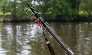 Gotowa do akcji wędka teleskopowa na spławik, z widokiem na spokojną wodę i zielone drzewa w tle.