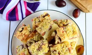 Pyszne ciasto drożdżowe ze śliwkami, posypane kruszonką, ułożone na talerzu. Obok kilka świeżych śliwek.