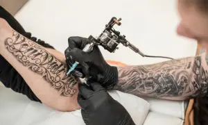 Artysta tworzy nowy tatuaż na przedramieniu klienta. Widać maszynkę do tatuażu i czarne rękawiczki.