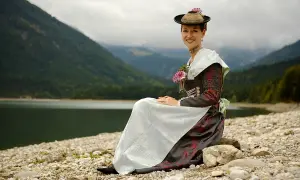 Frau in traditioneller Isarwinkler Tracht sitzt am Ufer eines Sees vor einer Bergkulisse.