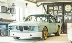 En grön BMW E9 i verkstad, redo för renovering. Guldiga fälgar och en imponerande bur syns tydligt.