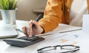 Osoba liczy na kalkulatorze, przygotowując się do zapłaty czynszu administracyjnego. Na biurku leżą dokumenty i okulary.