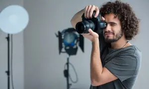 Mężczyzna z kręconymi włosami i aparatem w dłoniach, ćwiczący, jak zostać fotografem. W tle lampy studyjne.