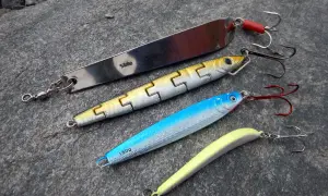 Vier Kunstköder für das Pilken vom Ufer liegen auf grauem Stein. Ein langer Silberpilker, ein gelb-silberner Wobbler, ein blauer Wobbler und ein gelber Gummifisch.