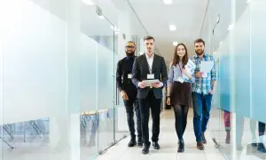 Fyra kollegor går genom en modern kontorskorridor. De ser ut att vara på väg till ett möte, kanske för att diskutera en affärsidé eller en potentiell finders fee.