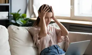 Kobieta z rękami na głowie siedzi na kanapie przed laptopem, zmagając się z prokrastynacją. Psychologia tego zjawiska jest złożona.