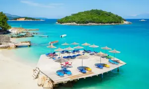 Albania jakie morze? Turkusowa woda, piaszczysta plaża, leżaki i parasole na drewnianym pomoście. Widok na zieloną wyspę.