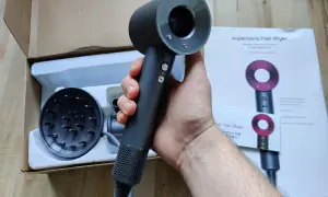 Dłoń trzymająca czarną suszarkę do włosów Dyson Supersonic obok pudełka i dyfuzora.