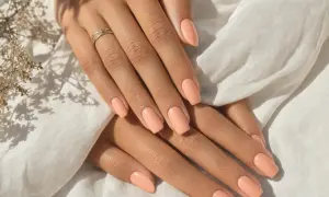 Mains aux ongles pêche, peau bronzée, bague fine. Douceur et élégance estivales.