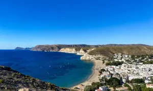 Vista panorámica de una **playa** con **agua amarga** de color azul intenso, un pueblo blanco y colinas áridas bajo un cielo despejado.
