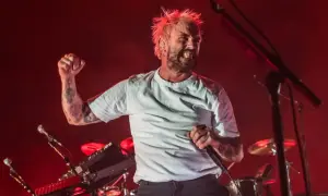A tattooed singer with spiky blonde hair belts into a microphone, fist raised, during a live performance. The idles setlist is in full swing.