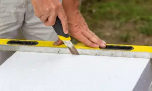 Ręce trzymają nóż, którym ciąć styropian, używając poziomicy jako prowadnicy.