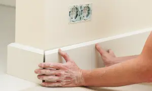 Ręce z pyłem gipsowym przyklejają białe listwy podłogowe do ściany. W tle widać gniazdka elektryczne.