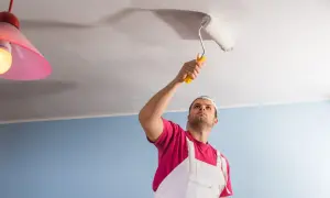Un homme en salopette blanche et t-shirt rose peint le plafond avec un rouleau. Il semble concentré sur son travail, donnant un nouveau sens à la peinture de plafond.