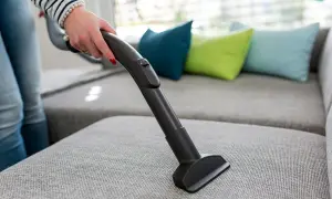En hand använder en soff dammsugare för att rengöra en grå soffa med färgglada kuddar i bakgrunden.