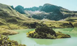 Grüne Hügel und ein türkisfarbener See laden zum Bergwandern in Bayern ein. Eine kleine Insel mit Bäumen liegt im Wasser.