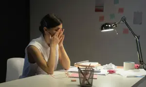 Zmęczona kobieta z rękami na twarzy, siedząca przy biurku po ustaniu zatrudnienia. Lampka biurkowa oświetla stos papierów.