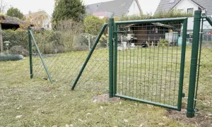 Ein grüner Maschendrahtzaun wird gespannt, mit einem Tor, das offen steht. Im Hintergrund ein Garten mit Häusern und einem Schuppen.