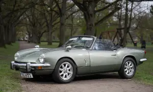 En ljusgrön Triumph Spitfire cabriolet parkerad på en grusväg i en allé.