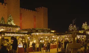 Mercado navideño bajo un castillo iluminado. Gente pasea entre puestos decorados con luces, mostrando como se celebra la navidad.