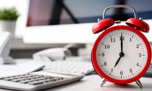 Czerwony budzik na biurku obok kalkulatora i komputera. Zastanawiasz się, ile godzin to 3/5 etatu? Czas pokaże!