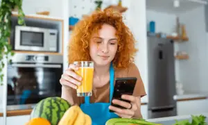 Rudowłosa kobieta z sokiem w dłoni szuka w telefonie informacji, co jeść na poprawę wzroku. Na blacie owoce i warzywa.