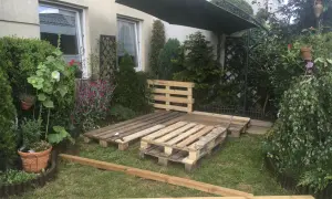 Taras z palet w trakcie budowy, otoczony zielenią i kwiatami. Pod dużym parasolem powstaje miejsce do relaksu.