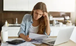 Zmartwiona kobieta analizuje dokumenty dotyczące sprzedaży mieszkania przed upływem 5 lat. Obok laptop, kalkulator i kubek.