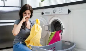 Donna con espressione disgustata annusa un asciugamano giallo, la lavatrice puzza. Cesto con panni colorati accanto.