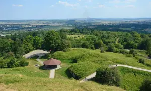 Zielony kopiec tatarski w Przemyślu z altaną i parkingiem. W oddali widać panoramę miasta i dymiące kominy.