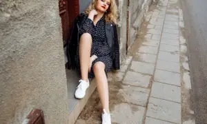 Kobieta w czarnej sukience w grochy i skórzanej kurtce, z białymi butami do czarnej sukienki, siedzi na progu.