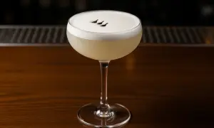 En elegant pisco sour med ett fluffigt skum och tre små mörka prickar på toppen, serverad i ett coupeglas på en träbar.