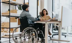 Mężczyzna na wózku inwalidzkim rozmawia z kobietą przy biurku. Pokazuje, że umiarkowany stopień niepełnosprawności a praca w ochronie to nie wykluczające się połączenie.