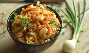 Aksamitne pad thai z krewetkami, posypane szczypiorkiem i orzeszkami, czeka w czarnej misce na drewnianym blacie. Obok leży cebulka.