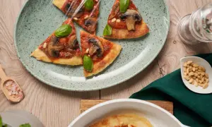 En tunn, krispig flatbread pizza med svamp och basilika, täckt med plastfolie. Pinjenötter och rosa salt ligger bredvid.
