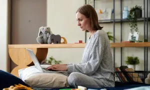 Kobieta w piżamie pisze na laptopie, przygotowując wniosek o urlop macierzyński. Na łóżku obok leżą zabawki.