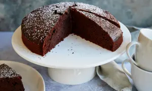 Ein saftiger veganer Schokokuchen, bestäubt mit Puderzucker, steht auf einem weißen Tortenständer. Daneben eine Tasse und ein Stück Kuchen auf einem Teller.