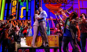 Szene aus einem Musical in Deutschland: Ein Paar umarmt sich, umgeben von jubelnden Darstellern vor einer bunten Kulisse mit Neonlichtern.