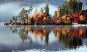 Ein idyllischer Urlaub im Oktober in Deutschland: Nebel steigt vom See auf, bunte Herbstbäume spiegeln sich im Wasser, eine grüne Ruderboot gleitet vorbei.